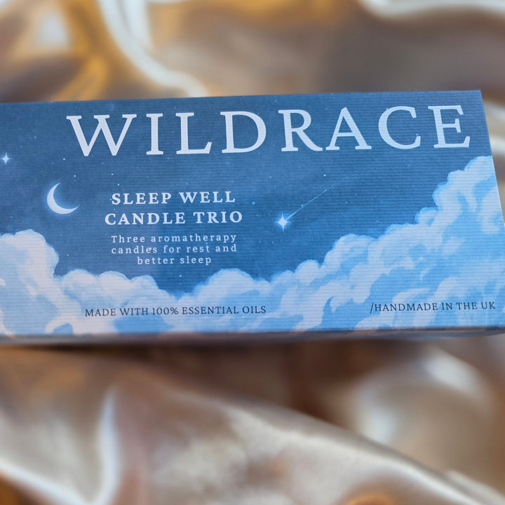 Wildrace 'Sleep Well' Candle Trio box, featuring a blue night sky with clouds and stars, resting on a draped beige satin fabric background.