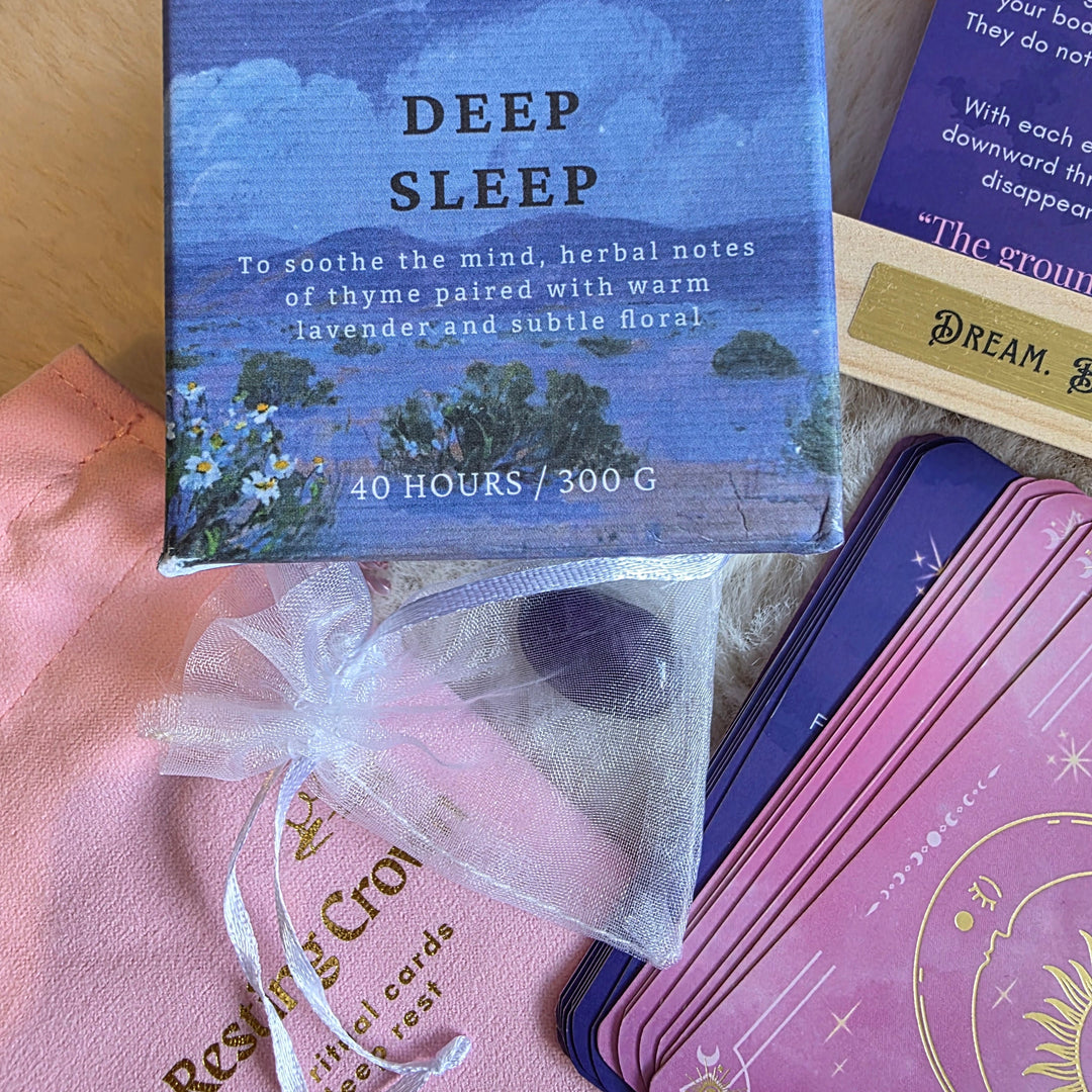 A Wildrace Scented candle  DEEP SLEEP is shown with a stack of pink Resting Crown sleep cards alongide its pink fabric pouch, a small clear drawstring bag containing a purple crystal,on a soft beige surface.