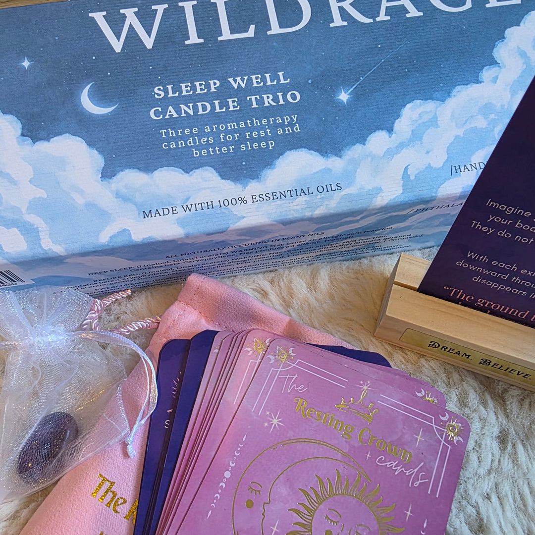 Wildrace Sleep Well Candle Trio set displayed on a white faux fur surface, including the blue and white scented candle box, a wooden card stand with a tarot card, a pink and purple card deck, and a small crystal bag.