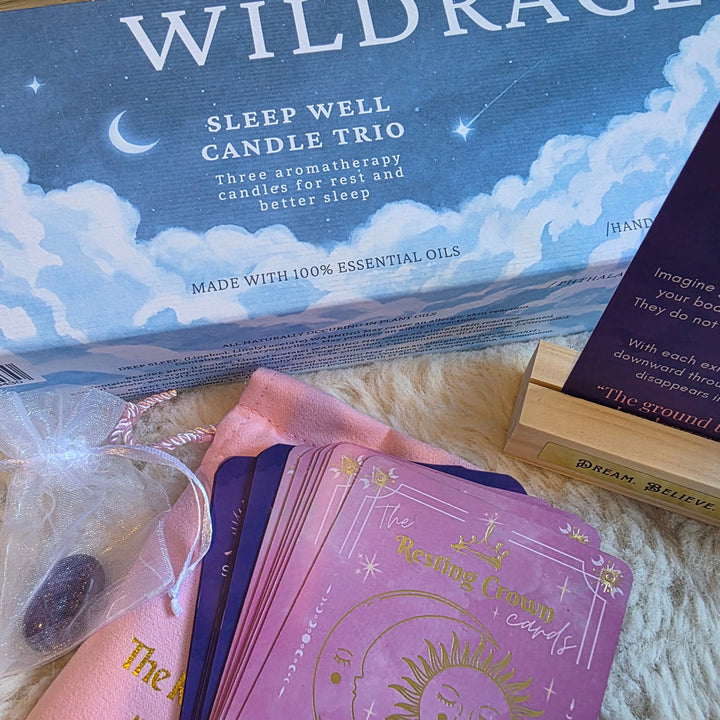 Wildrace Sleep Well Candle Trio set displayed on a white faux fur surface, including the blue and white scented candle box, a wooden card stand with a tarot card, a pink and purple card deck, and a small crystal bag.