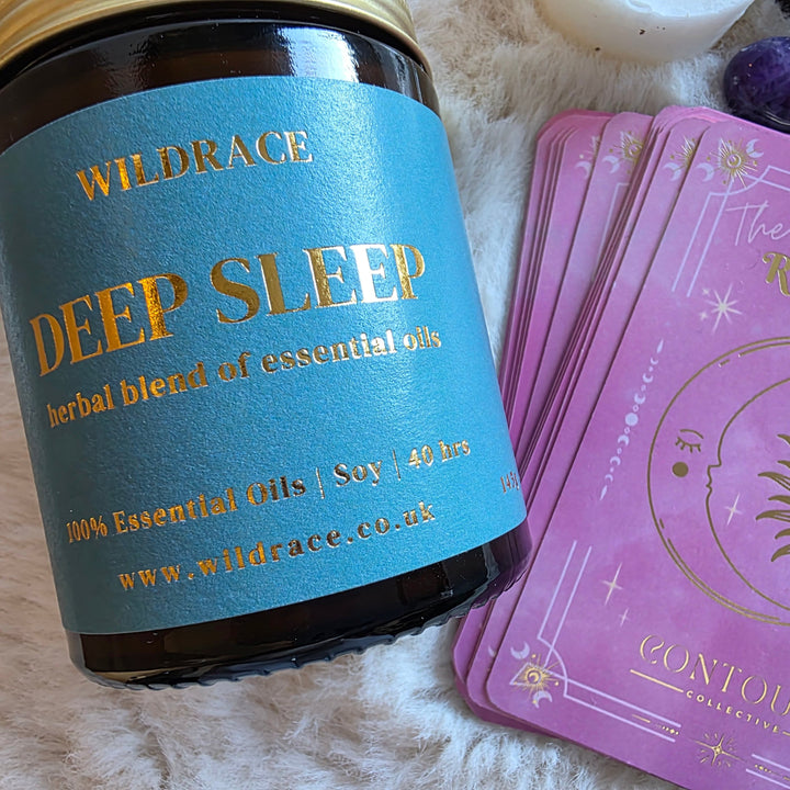 A Wildrace Deep Sleep essential oil scented candle lies on a white faux fur surface next to a stack of pink Resting Crown Sleep cards with gold celestial artwork and a purple crystal.
