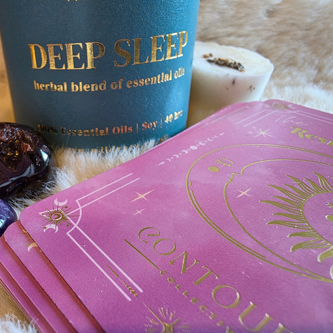 A pink Contoura Collection Resting Crown sleep cards deck with gold foil celestial designs, shown next to a Wildrace Deep Sleep  essential oil scented candle, crystals, and a white wax melt on a white faux fur surface.