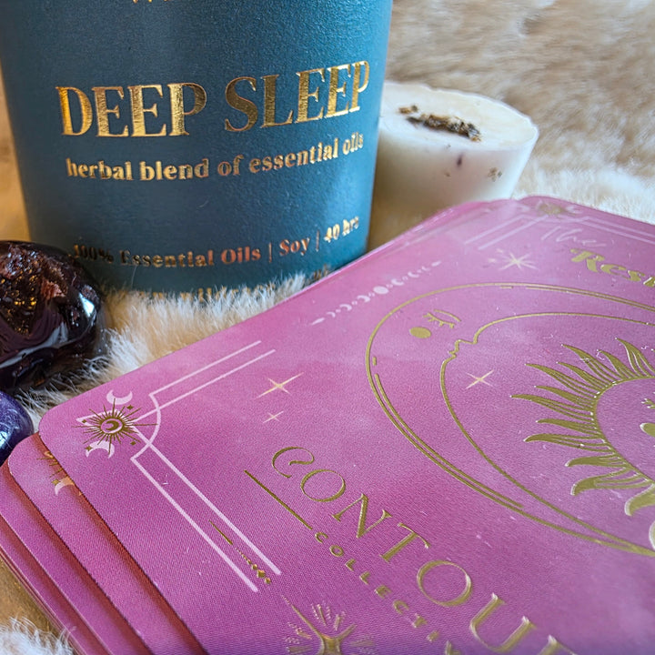 A pink Contoura Collection Resting Crown sleep cards deck with gold foil celestial designs, shown next to a Wildrace Deep Sleep  essential oil scented candle, crystals, and a white wax melt on a white faux fur surface.
