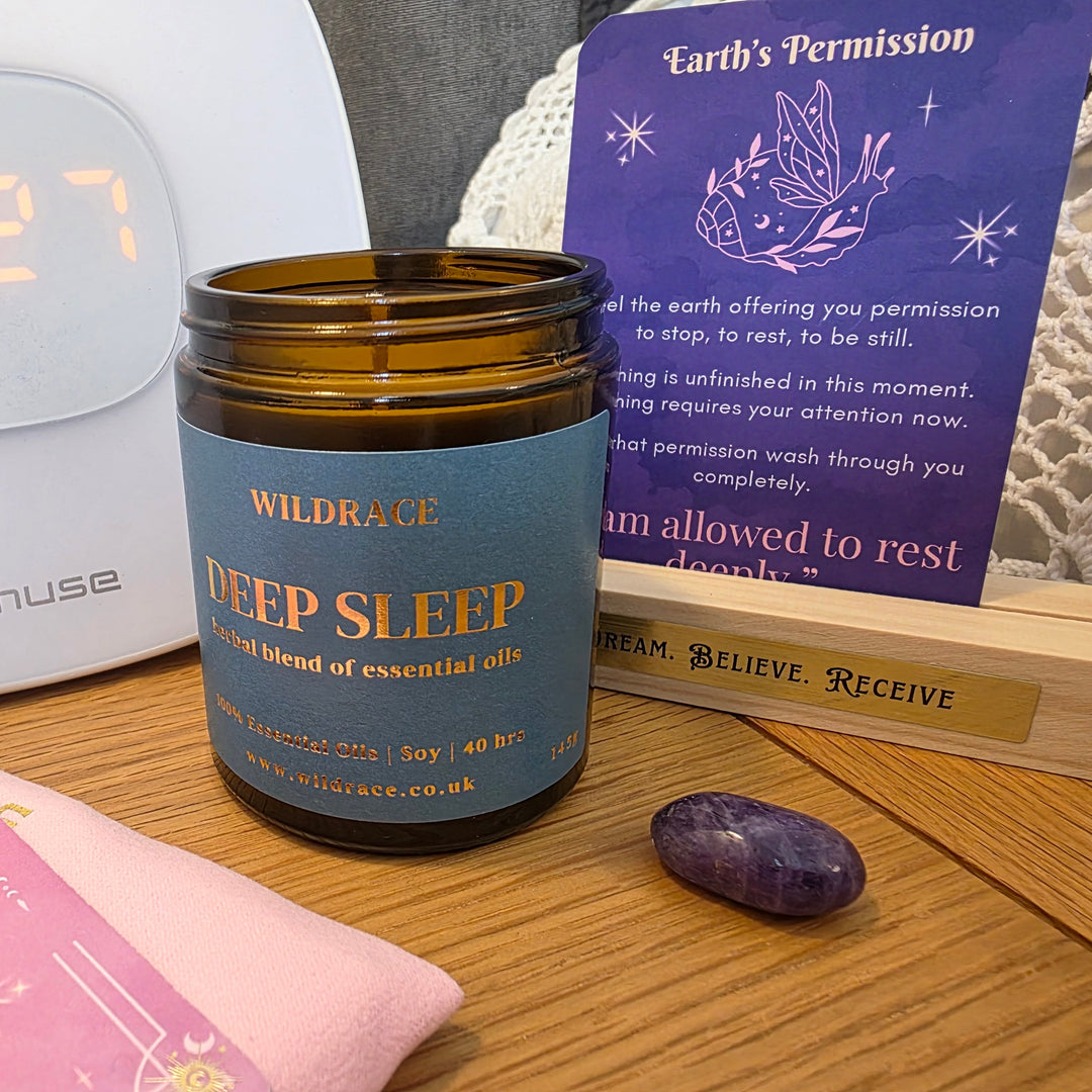 An amber glass jar of Wildraces Deep Sleep essential oil scented candle on a wooden table, a deck of Resting Crown Sleep cards to the side and a purple amethyst crystal, a digital clock, and a purple Resting Crown sleep deck card in a Wooden stand.