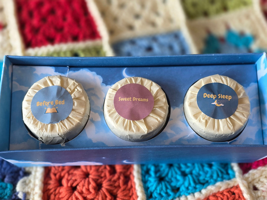 A blue gift box containing three wrapped scented candles, photographed close up on a colorful crocheted blanket background. The candles are labled 'Before Bed', 'Sweet Dreams' and 'Deep Sleep'.