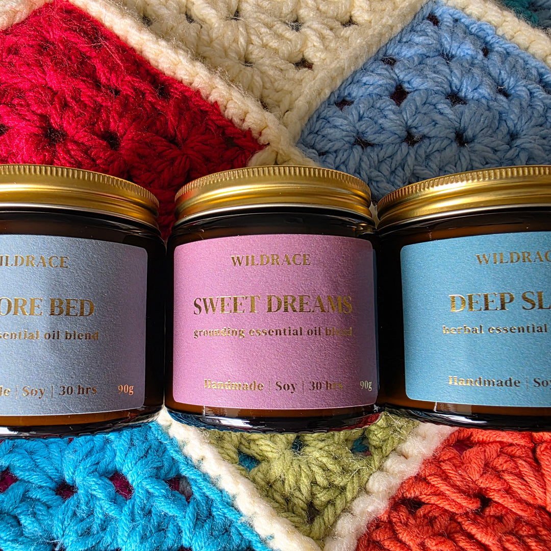Three amber glass jars of Wildrace essential oil scented candles with gold lids are lined up on a colorful crocheted blanket. The labels read 'WILDRACE BEFORE BED', 'WILDRACE SWEET DREAMS', and 'WILDRACE DEEP SLEEP'.