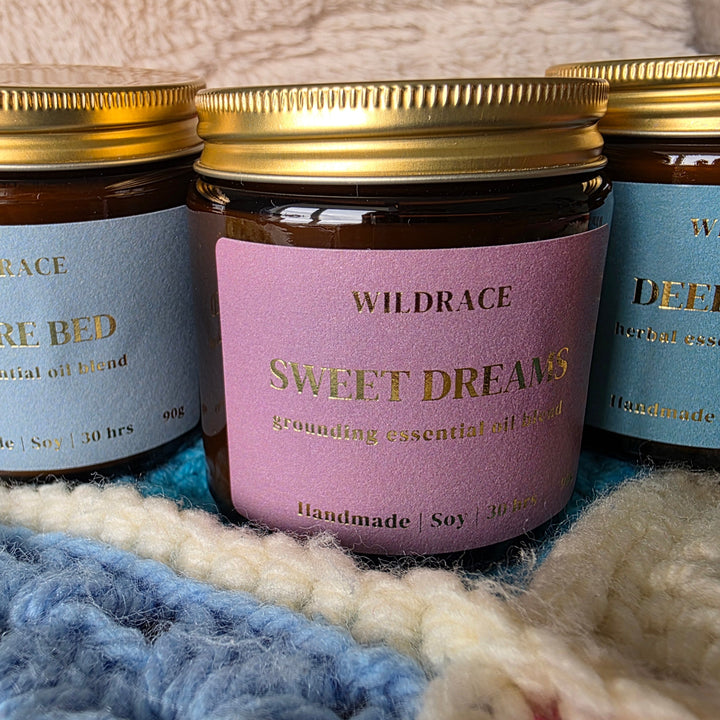 Three amber glass jars of Wildrace essential oil candles with gold lids, labeled Before Bed, Sweet Dreams, and Deep Sleep, arranged on a blue and cream knit blanket.