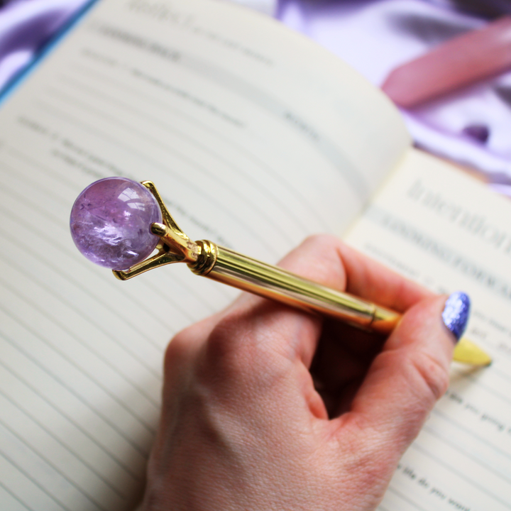A person journalling in a gratitude journal holding an Amethyst crystal ball pen