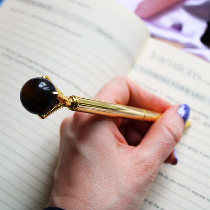 Person writing in a gratitude journal holding a Tigers Eye crystal ball pen