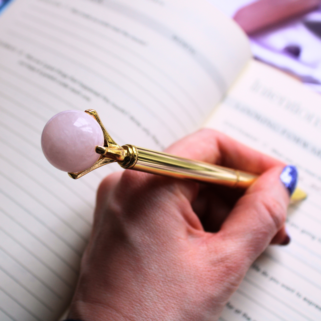 A person writing in a gratitude journal holding a Rose Quartz ball pen