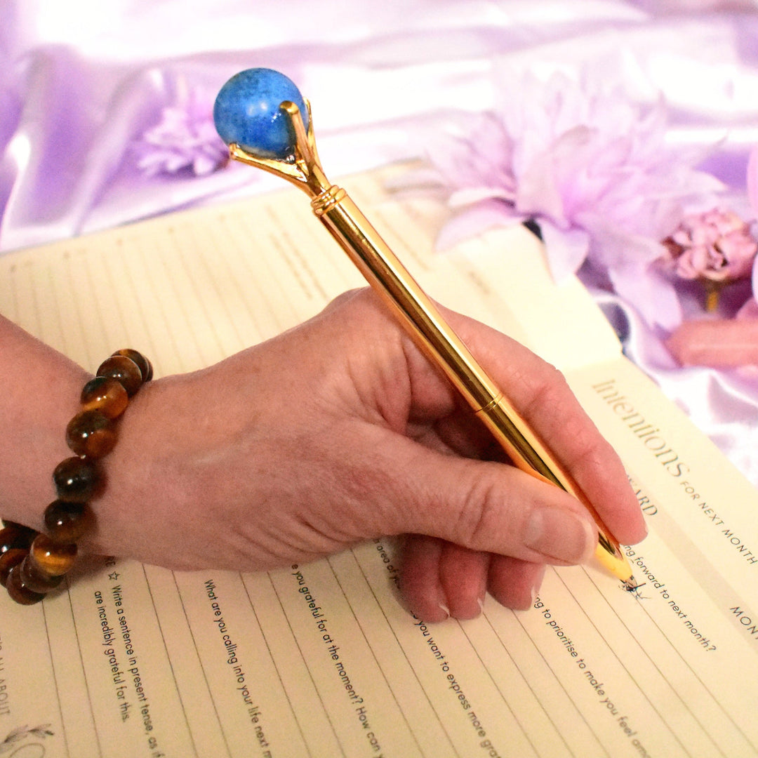Hand holding a gold pen with a blue gemstone on a piece of paper, with a decorative background.