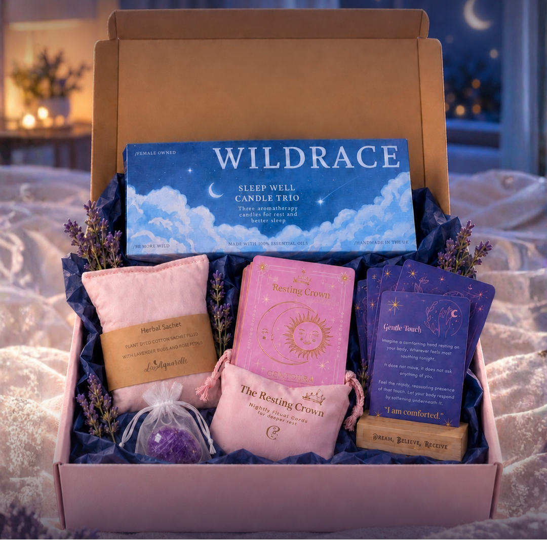 A gift box set labeled 'The Sleep Trio Gift Box' shown open with three themed sleep items inside: a blue 'Wildrace Sleep Well Candle Trio' box, a pink lavender sachet, a set of Resting Crown Sleep cards, pink velvet storage bag and wooden stand, as well as an purple amethyst crystal. The scene is styled in a bedroom with a blurred nightstand and moonlit background.