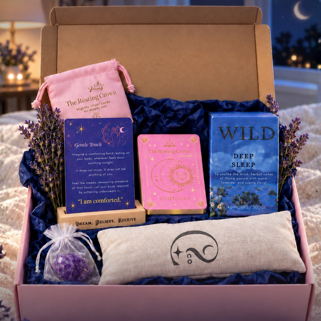 An open gift box filled with sleep-themed items, including a lavender sachet, a cream  lavender eye pillow with a Contoura logo, a Sleep card in a wooden stand, a deck of Sleep cards, a Wildrace scented candle 'DEEP SLEEP,' and a pink card pouch. The box is set on a bed with a nightstand and lamp in the background.